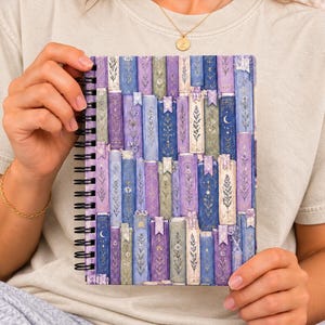 May include: A spiral-bound notebook featuring a cover illustration of stacked books in shades of purple, blue, and green. The book spines are decorated with gold botanical patterns, stars, and crescent moons, creating a detailed, layered visual design.