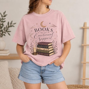 Pode incluir: Uma t-shirt rosa claro com uma estampa gráfica de uma pilha de livros, uma vela acesa e uma lua crescente. O texto "BOOKS ARE MY Emotional Support" está escrito numa mistura de fontes serifadas e manuscritas, rodeado por estrelas e folhagens.
