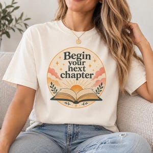 May include: A cream-coloured short-sleeve t-shirt featuring a circular graphic with the text "Begin your next chapter" in dark lettering. The design includes an open book, a rising sun, pink clouds, and small star accents centred on the chest.