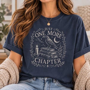 Pode incluir: Uma t-shirt de manga curta azul-marinho com uma estampa gráfica de um corvo pousado numa pilha de livros ao lado de uma vela acesa e uma lua crescente. O texto "JUST ONE MORE CHAPTER" está impresso numa moldura circular com ramos botânicos.