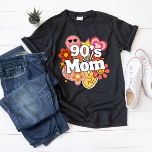 Op de afbeelding: Zwart T-shirt met de tekst "90's Mom" in retrostijl, omringd door kleurrijke graphics. Het shirt wordt getoond met een blauwe jeans en witte sneakers.
