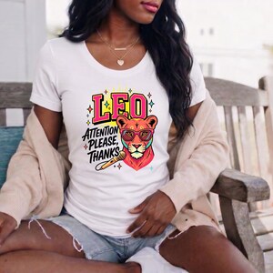 May include: A white short-sleeved t-shirt featuring a graphic of a lion wearing pink glasses, a microphone, and the text LEO ATTENTION PLEASE, THANKS. The design includes colorful star accents and bold lettering in a vibrant, multi-colored style.