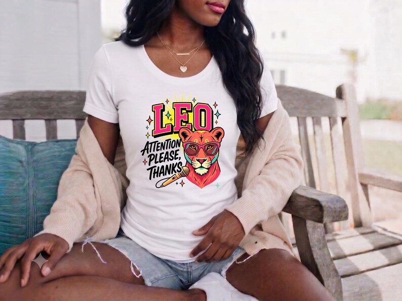 May include: A white short-sleeved t-shirt featuring a graphic of a lion wearing pink glasses, a microphone, and the text LEO ATTENTION PLEASE, THANKS. The design includes colorful star accents and bold lettering in a vibrant, multi-colored style.