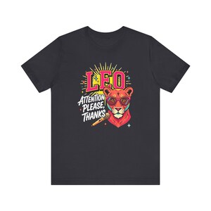 May include: A dark gray short-sleeve t-shirt featuring a colorful graphic of a lion wearing pink sunglasses. The design includes the text LEO and ATTENTION PLEASE, THANKS, along with a microphone illustration and small star accents in yellow and blue.
