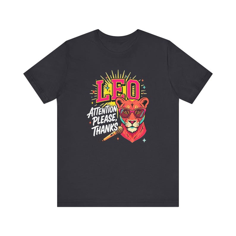 May include: A dark gray short-sleeve t-shirt featuring a colorful graphic of a lion wearing pink sunglasses. The design includes the text LEO and ATTENTION PLEASE, THANKS, along with a microphone illustration and small star accents in yellow and blue.