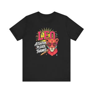 May include: A black t-shirt featuring a graphic of a lion wearing pink glasses. The lion is positioned next to a microphone. Above the lion, the text reads LEO in pink, and below that, the text reads ATTENTION PLEASE, THANKS in white letters.