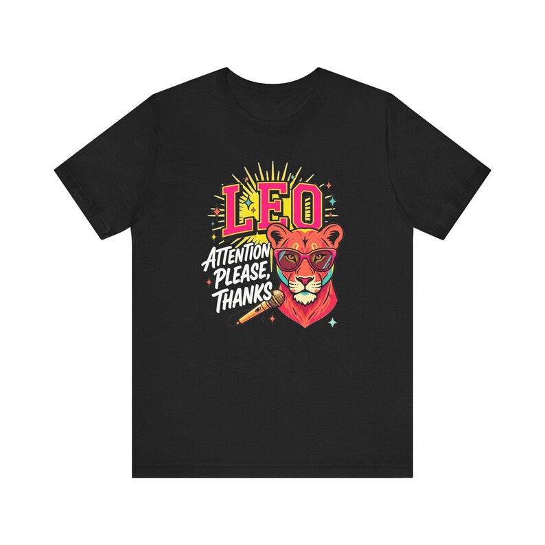 May include: A black t-shirt featuring a graphic of a lion wearing pink glasses. The lion is positioned next to a microphone. Above the lion, the text reads LEO in pink, and below that, the text reads ATTENTION PLEASE, THANKS in white letters.