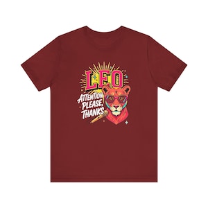 May include: A maroon short-sleeve t-shirt featuring a graphic of a lion wearing pink glasses. The design includes the text LEO and ATTENTION PLEASE, THANKS, along with a microphone illustration, yellow sunburst rays, and small star accents.