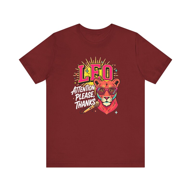 May include: A maroon short-sleeve t-shirt featuring a graphic of a lion wearing pink glasses. The design includes the text LEO and ATTENTION PLEASE, THANKS, along with a microphone illustration, yellow sunburst rays, and small star accents.