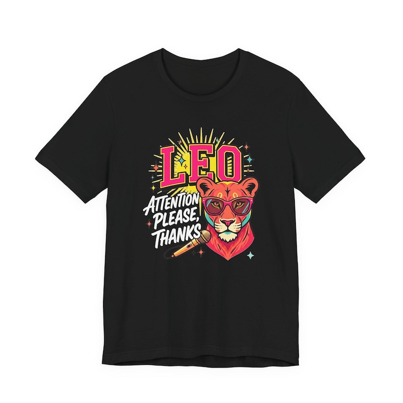 May include: A black t-shirt featuring a colorful graphic of a lion wearing pink-rimmed glasses. The design includes the text LEO and ATTENTION PLEASE, THANKS, along with a microphone illustration, surrounded by small star-shaped accents.