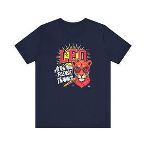 May include: A navy blue t-shirt featuring a graphic of a lion wearing pink-rimmed sunglasses. The lion is positioned next to a microphone. Above the lion, the text reads LEO in pink, and below, the text reads ATTENTION PLEASE, THANKS in white.