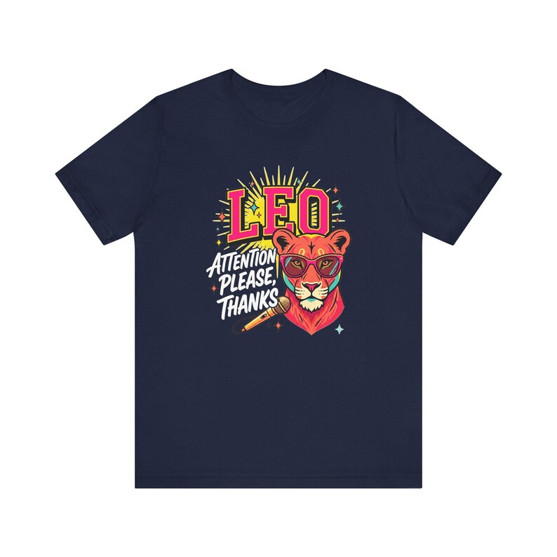 May include: A navy blue t-shirt featuring a graphic of a lion wearing pink-rimmed sunglasses. The lion is positioned next to a microphone. Above the lion, the text reads LEO in pink, and below, the text reads ATTENTION PLEASE, THANKS in white.