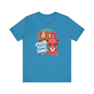May include: A bright blue short-sleeve t-shirt featuring a graphic of a lion wearing pink-rimmed sunglasses. The design includes the text LEO and ATTENTION PLEASE, THANKS, along with a microphone illustration and yellow sunburst accents.
