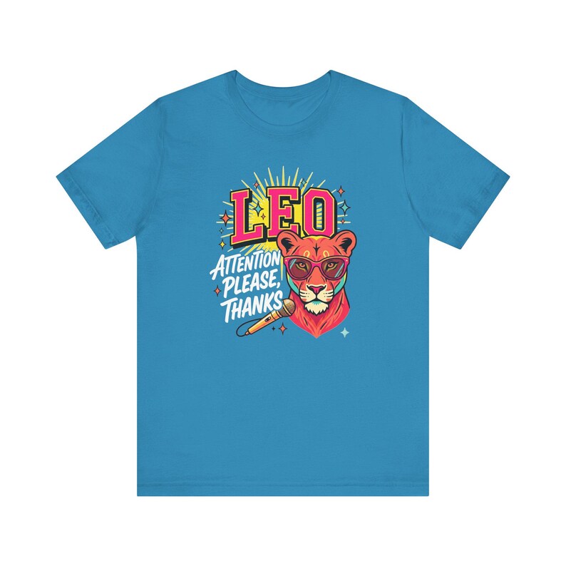 May include: A bright blue short-sleeve t-shirt featuring a graphic of a lion wearing pink-rimmed sunglasses. The design includes the text LEO and ATTENTION PLEASE, THANKS, along with a microphone illustration and yellow sunburst accents.