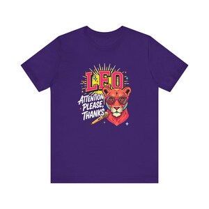 May include: A purple short-sleeve t-shirt featuring a graphic of a lion wearing pink glasses. The design includes the text LEO and ATTENTION PLEASE, THANKS, along with a microphone illustration, yellow sunburst rays, and small colorful star accents.