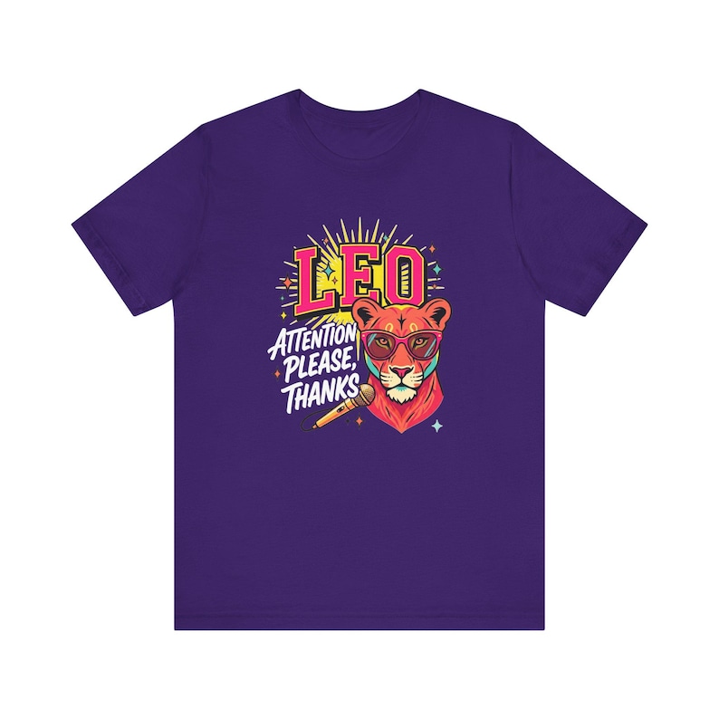 May include: A purple short-sleeve t-shirt featuring a graphic of a lion wearing pink glasses. The design includes the text LEO and ATTENTION PLEASE, THANKS, along with a microphone illustration, yellow sunburst rays, and small colorful star accents.