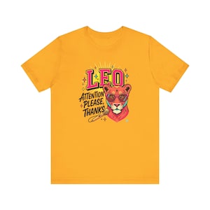 May include: A golden yellow short-sleeve t-shirt featuring a graphic of a lion wearing pink sunglasses. The text reads LEO and ATTENTION PLEASE, THANKS, accompanied by a microphone illustration and small star accents in pink, blue, and yellow.