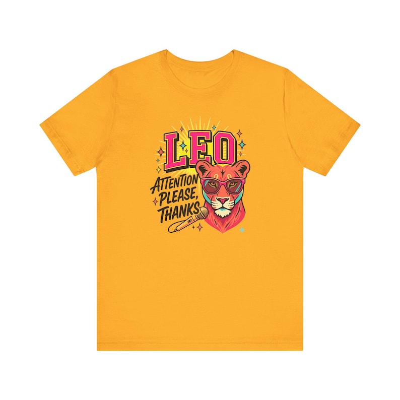 May include: A golden yellow short-sleeve t-shirt featuring a graphic of a lion wearing pink sunglasses. The text reads LEO and ATTENTION PLEASE, THANKS, accompanied by a microphone illustration and small star accents in pink, blue, and yellow.
