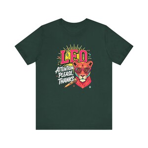 May include: A dark green t-shirt featuring a graphic of a lion wearing pink sunglasses. The text reads LEO and ATTENTION PLEASE, THANKS. A microphone is positioned below the lion's chin, surrounded by yellow sunburst rays and small colorful stars.