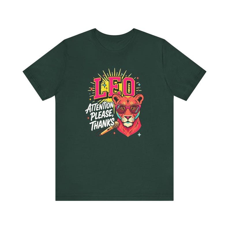 May include: A dark green t-shirt featuring a graphic of a lion wearing pink sunglasses. The text reads LEO and ATTENTION PLEASE, THANKS. A microphone is positioned below the lion's chin, surrounded by yellow sunburst rays and small colorful stars.