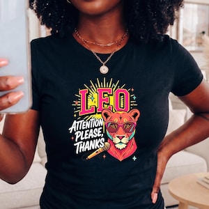 May include: A black short-sleeved t-shirt featuring a colorful graphic of a lion wearing pink-rimmed glasses. The design includes the text LEO and ATTENTION PLEASE, THANKS, along with a microphone illustration and yellow starburst accents.