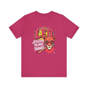 May include: A dark pink short-sleeve t-shirt featuring a graphic of a lion wearing pink sunglasses. The text LEO and ATTENTION PLEASE, THANKS is printed above and beside the lion, accompanied by a microphone and small star-shaped accents.