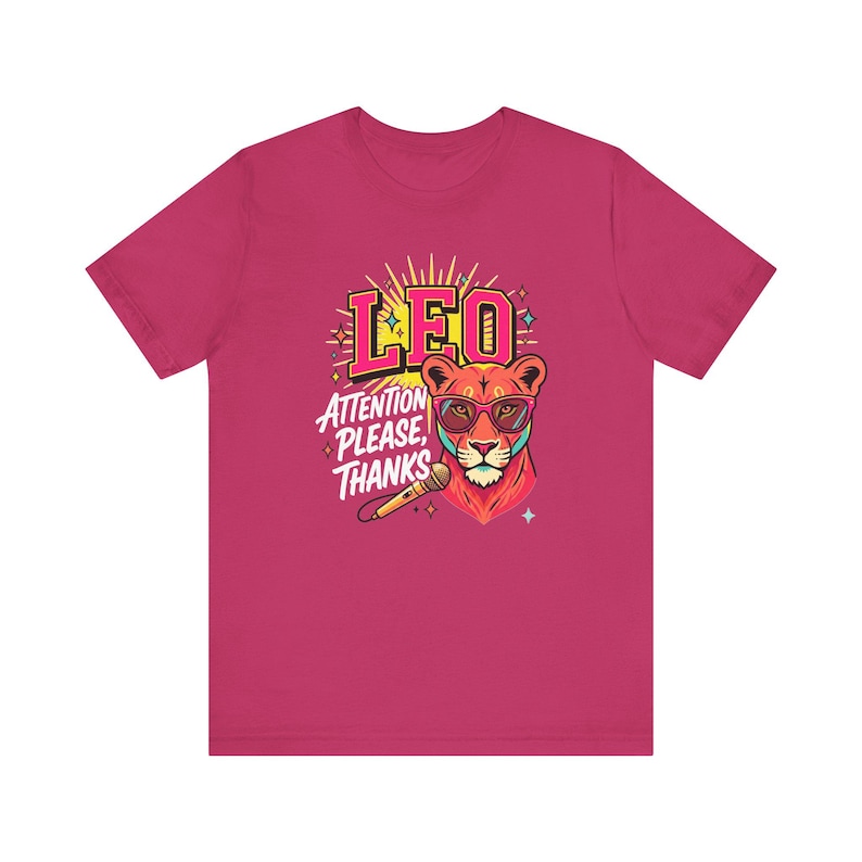 May include: A dark pink short-sleeve t-shirt featuring a graphic of a lion wearing pink sunglasses. The text LEO and ATTENTION PLEASE, THANKS is printed above and beside the lion, accompanied by a microphone and small star-shaped accents.