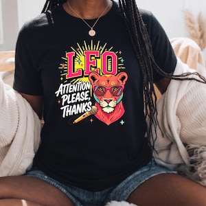 May include: A black t-shirt featuring a graphic of a lion wearing pink-rimmed glasses, holding a microphone, with the text LEO ATTENTION PLEASE, THANKS in pink and white lettering, surrounded by yellow sunburst rays and small star accents.