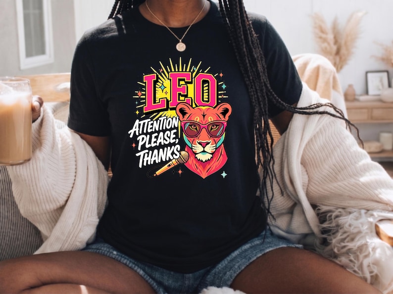 May include: A black t-shirt featuring a graphic of a lion wearing pink-rimmed glasses, holding a microphone, with the text LEO ATTENTION PLEASE, THANKS in pink and white lettering, surrounded by yellow sunburst rays and small star accents.