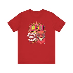 May include: A red short-sleeve t-shirt featuring a graphic of a lion wearing pink glasses. The text reads LEO and ATTENTION PLEASE, THANKS. A microphone is positioned below the lion's head, surrounded by yellow sunburst rays and small stars.