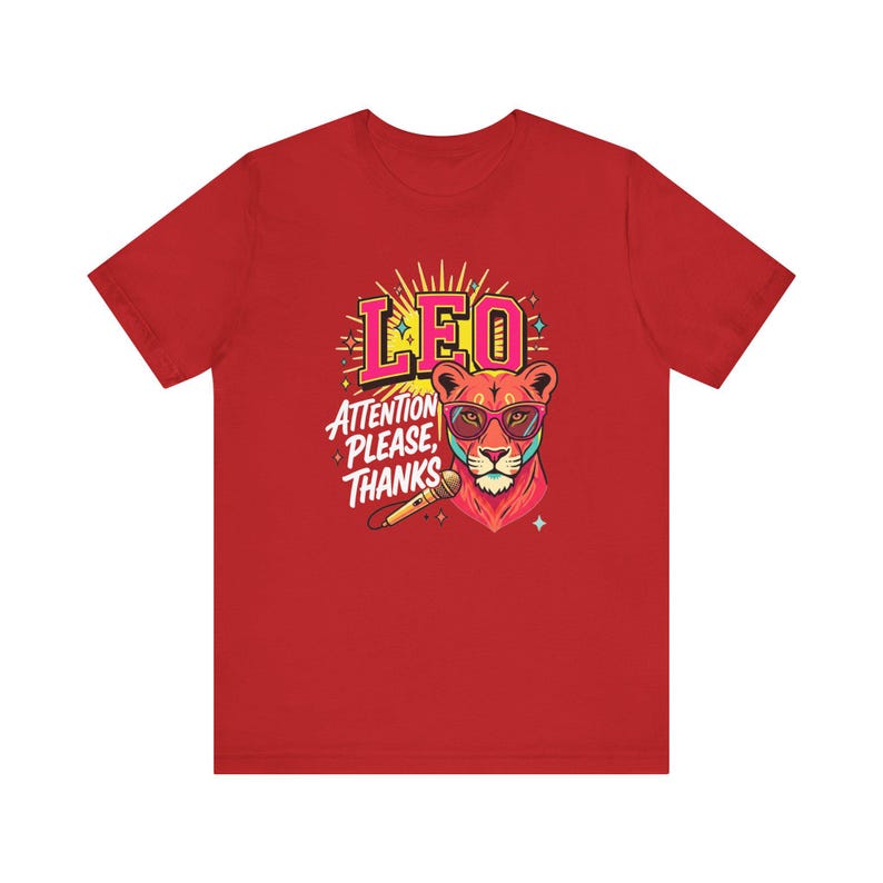 May include: A red short-sleeve t-shirt featuring a graphic of a lion wearing pink glasses. The text reads LEO and ATTENTION PLEASE, THANKS. A microphone is positioned below the lion's head, surrounded by yellow sunburst rays and small stars.