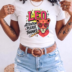 May include: A white short-sleeve t-shirt featuring a graphic of a lion wearing pink-rimmed glasses, a microphone, and the text LEO ATTENTION PLEASE, THANKS in bold pink and black lettering, accented with colorful star shapes and yellow sunburst rays.