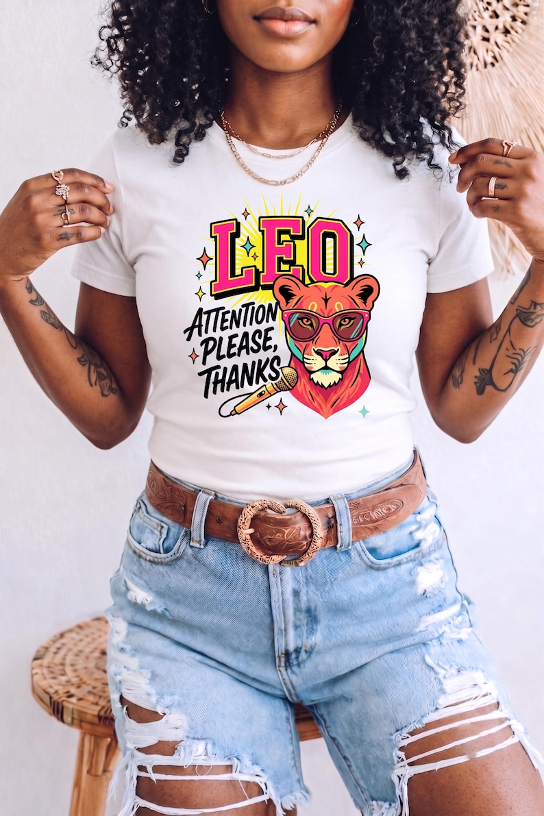 May include: A white short-sleeve t-shirt featuring a graphic of a lion wearing pink-rimmed glasses, a microphone, and the text LEO ATTENTION PLEASE, THANKS in bold pink and black lettering, accented with colorful star shapes and yellow sunburst rays.