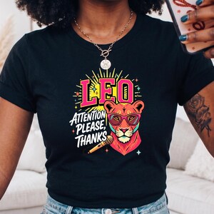 May include: A black short-sleeve t-shirt featuring a colorful graphic of a lion wearing pink sunglasses. The design includes the text LEO and ATTENTION PLEASE, THANKS, along with a microphone illustration, yellow sunburst rays, and small star accents.
