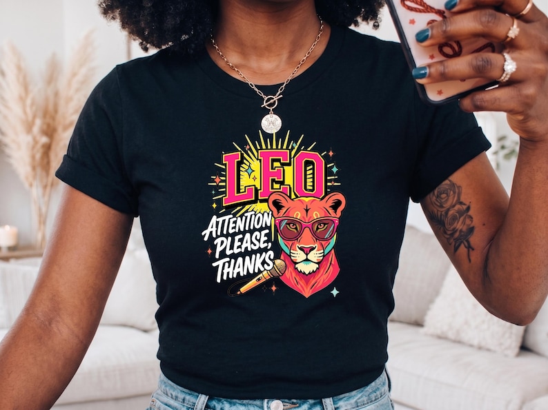 May include: A black short-sleeve t-shirt featuring a colorful graphic of a lion wearing pink sunglasses. The design includes the text LEO and ATTENTION PLEASE, THANKS, along with a microphone illustration, yellow sunburst rays, and small star accents.