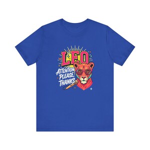 May include: A royal blue short-sleeve t-shirt featuring a graphic of a lion wearing pink glasses. The design includes the text LEO and ATTENTION PLEASE, THANKS, along with a microphone illustration and yellow sunburst rays behind the lion head.