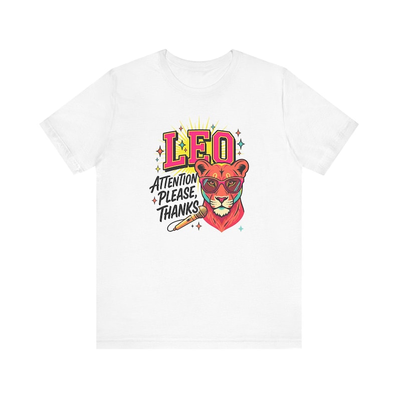 May include: A white short-sleeve t-shirt featuring a graphic of a lion wearing pink sunglasses and holding a microphone. The text LEO and ATTENTION PLEASE, THANKS is printed in bold colors above and beside the lion, surrounded by small star icons.