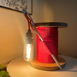 May include: A table lamp featuring a wooden spool wrapped in bright red cord, with a white knitting needle protruding through the center. A single Edison-style light bulb hangs from a red cord attached to the needle, emitting a warm glow.