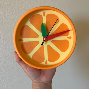 May include: A round wall clock featuring a vibrant orange citrus slice design on the face. The clock has a bright orange frame, a pale yellow background with orange segments, and three distinct clock hands in green, red, and black.