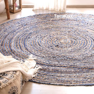 May include: A round area rug featuring a coiled design made from braided blue fabric strips and natural tan jute fibers. The rug displays a concentric circular pattern throughout its surface and includes the text TerraKnot printed in white.