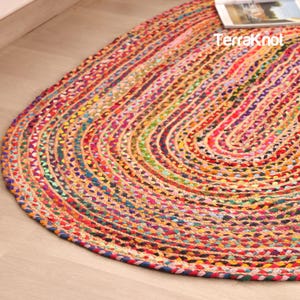 Op de afbeelding: Ovaal gevlochten tapijt met een levendige mix van kleuren, waaronder rood, oranje, geel, blauw en groen. Het tapijt is gemaakt van geweven stof en heeft de tekst "TerraKnot" erop. Het tapijt ligt op een lichtgekleurde houten vloer.