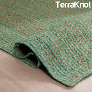 Puede incluir: Una alfombra rectangular enrollada con una textura tejida. La alfombra es de color verde vibrante con rayas color canela que corren a lo largo. El borde de la alfombra está cuidadosamente terminado, y la marca "TerraKnot" es visible en la esquina superior derecha.