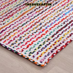 May include: A multi-coloured braided rug featuring a white base with vertical stripes in shades of red, orange, yellow, blue, and purple. The rug has a textured finish and is made from woven material. The text "THERUGSARTIN" is visible at the top.