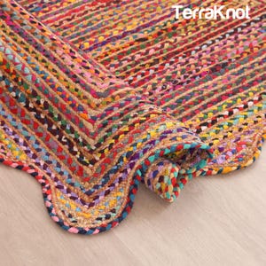 Op de afbeelding: Een rechthoekig vloerkleed met een gevlochten constructie en een veelkleurig patroon van rode, blauwe, gele, groene en paarse stoffen stroken, verweven met natuurlijke beige vezels. De merknaam TerraKnot staat in het wit in de hoek.