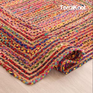 Könnte beinhalten: Ein rechteckiger geflochtener Teppich mit einem mehrfarbigen Muster aus ineinander verschlungenen roten, blauen, gelben und grünen Fasern. Der Teppich hat eine dicke, strukturierte Oberfläche mit einer sichtbaren Ecke. Der Text TerraKnot steht in Weiß.