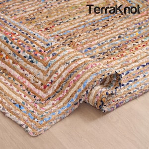 May include: A rectangular area rug with a braided design. The rug features a mix of natural jute fibers and colorful fabric strips in shades of blue, pink, and white. The rug is partially rolled up, revealing its texture and construction. The text "Terraknot" is visible.