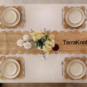 Puede incluir: Una mesa de comedor con mantel blanco, cuatro manteles individuales redondos de yute trenzado con bordes festoneados y un camino de mesa de yute a juego con el texto TerraKnot. Cuatro platos blancos, cubiertos, vasos y un centro floral.