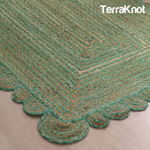 May include: A close-up of a jute rug with a scalloped edge. The rug features a geometric design with alternating stripes of green and tan. The text "TerraKnot" is in the upper right corner.