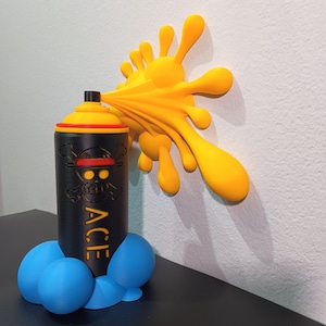 Può includere: Una bomboletta spray nera e gialla con un design a teschio e ossa incrociate, la parola "ACE" e un effetto di spruzzo di vernice gialla. La base è blu. L'opera d'arte è esposta su una superficie nera contro un muro bianco.