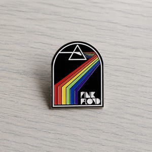 Puede incluir: Pin de esmalte negro con el diseño del arte del álbum de Pink Floyd. Presenta un triángulo blanco, un espectro de arcoíris y el nombre de la banda en letras blancas. El pin tiene un borde plateado y forma rectangular.